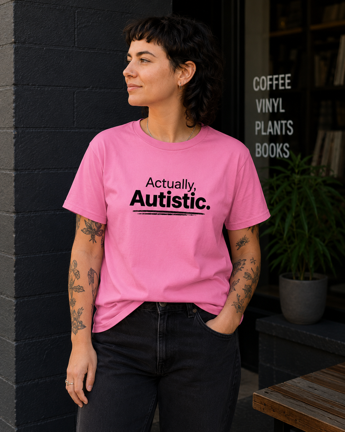 A woman with short dark hair and tattoos stands in front of a dark brick wall and storefront window, wearing a bright pink T-shirt with the black printed text 'Actually, Autistic.' and black jeans.