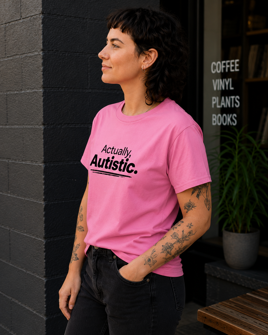 A woman wearing a pink graphic t-shirt with the text 'Actually Autistic.' printed in black, standing outdoors next to a dark brick wall and a window with the words 'COFFEE VINYL PLANTS BOOKS'.