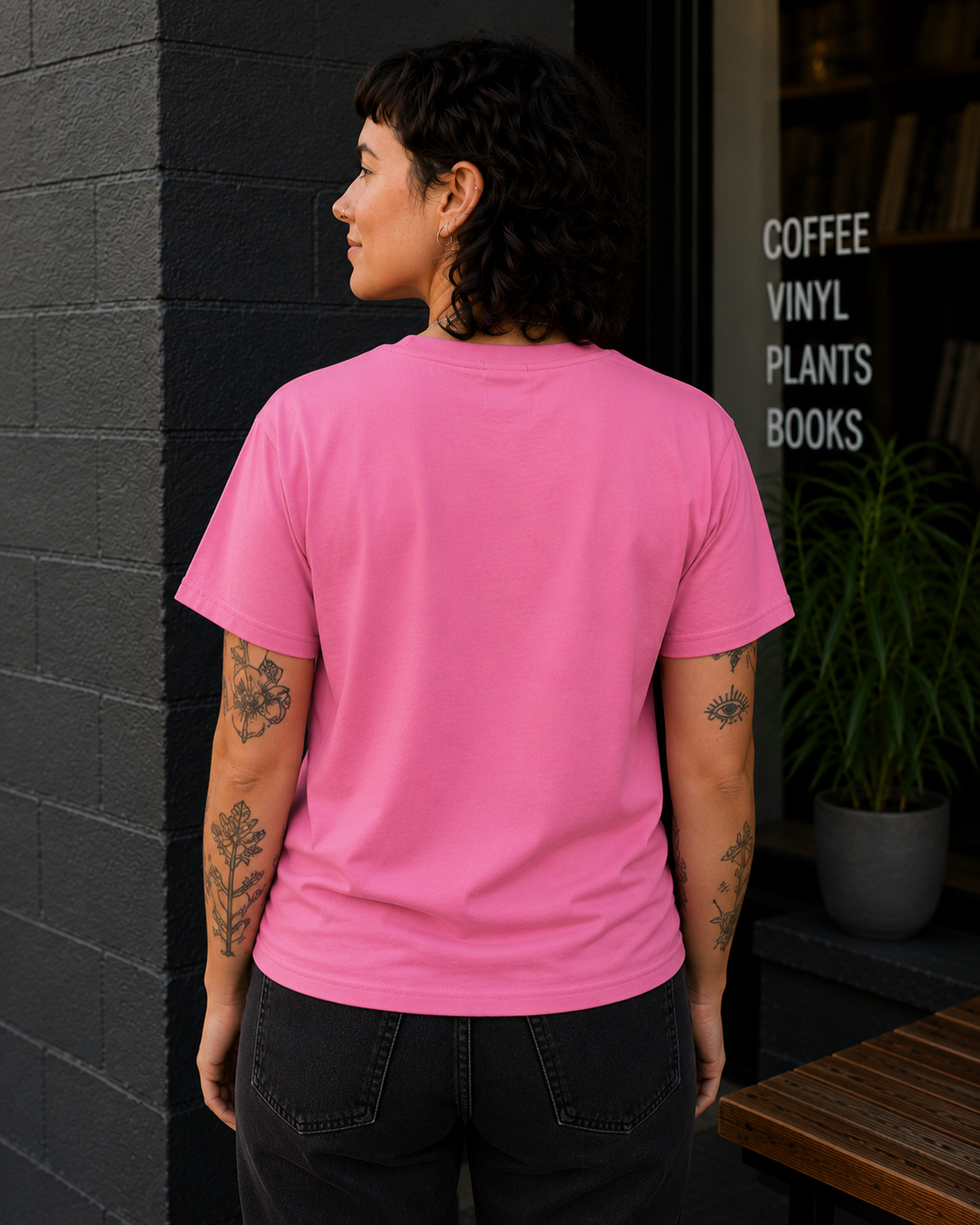 A person standing outdoors in front of a storefront, shown from the back wearing a bright pink short-sleeve T-shirt and dark jeans. The background includes a dark brick wall, a window with white text reading 'COFFEE VINYL PLANTS BOOKS,' and a potted plant.