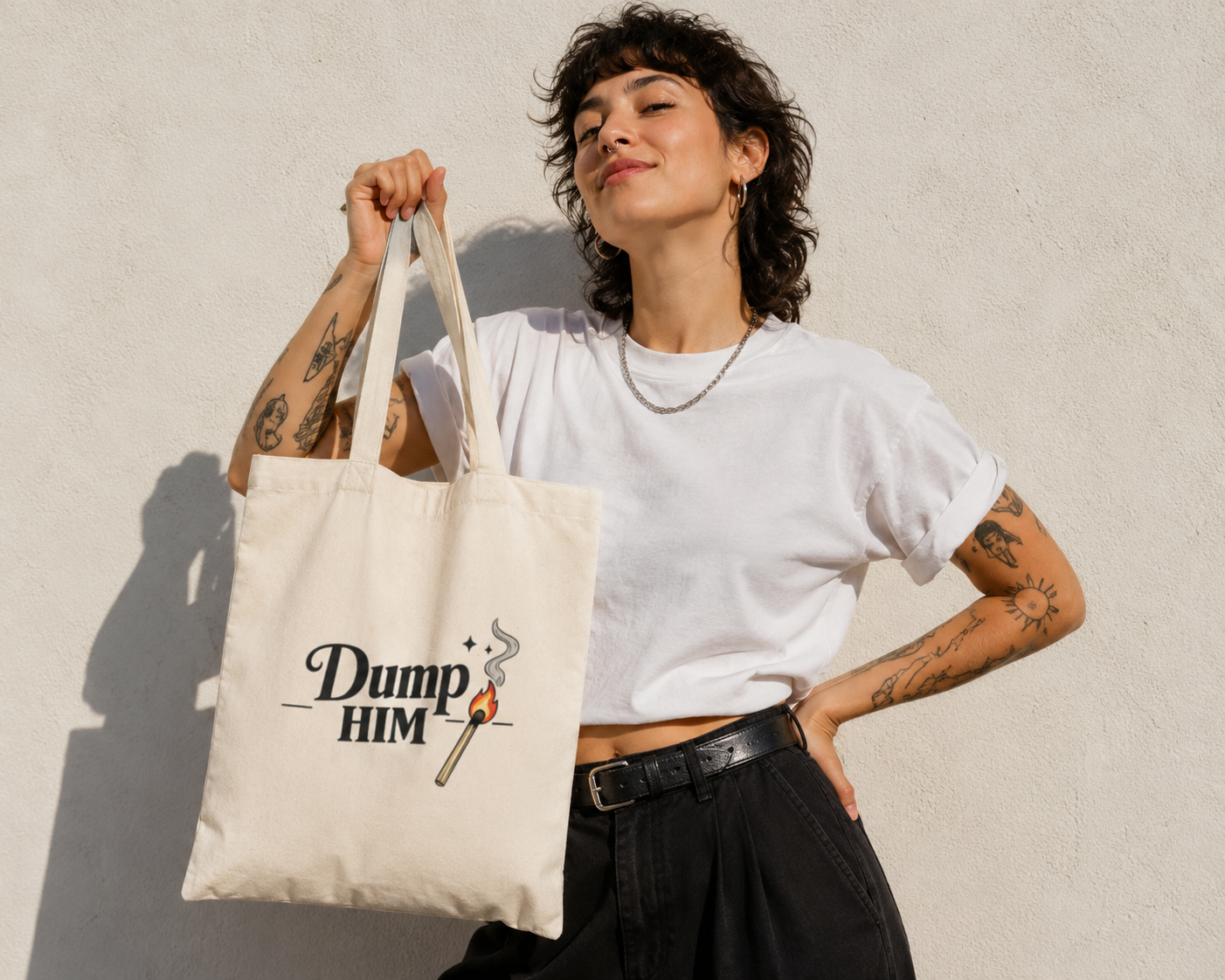 A woman with short curly dark hair and tattoos holds up a cream canvas tote bag with the black printed text 'Dump HIM' and a small flame graphic, wearing a white t-shirt and black pants against a textured light gray wall.