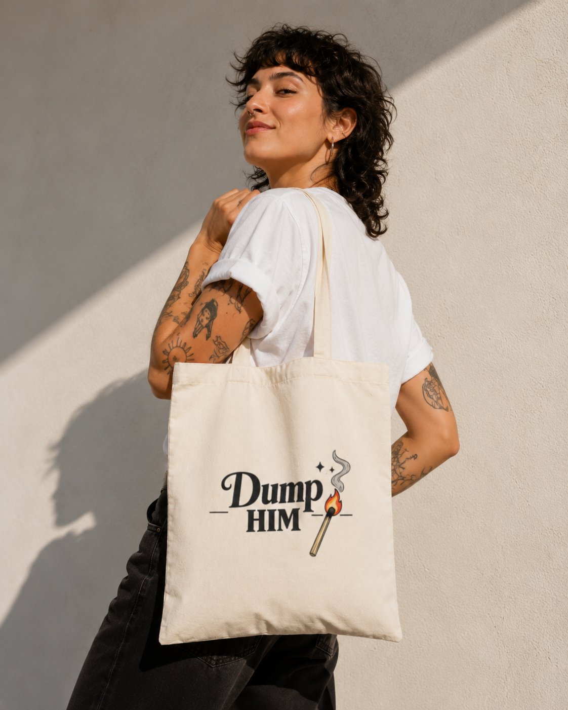 A woman with short curly dark hair, wearing a white t-shirt and black pants, carries a cream canvas tote bag over her shoulder. The bag features the text 'Dump HIM' in black script with a graphic of a burning match.