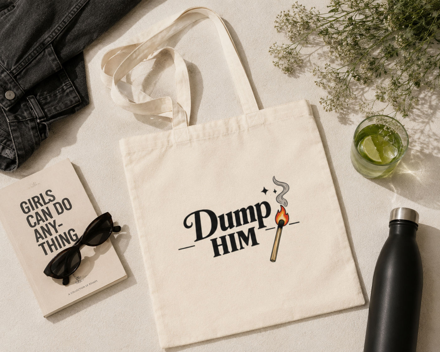 An off-white canvas tote bag with the text 'Dump HIM' and a match graphic printed on the front, styled in a flat-lay arrangement with a book, sunglasses, denim, a drink, and a water bottle on a light beige surface.