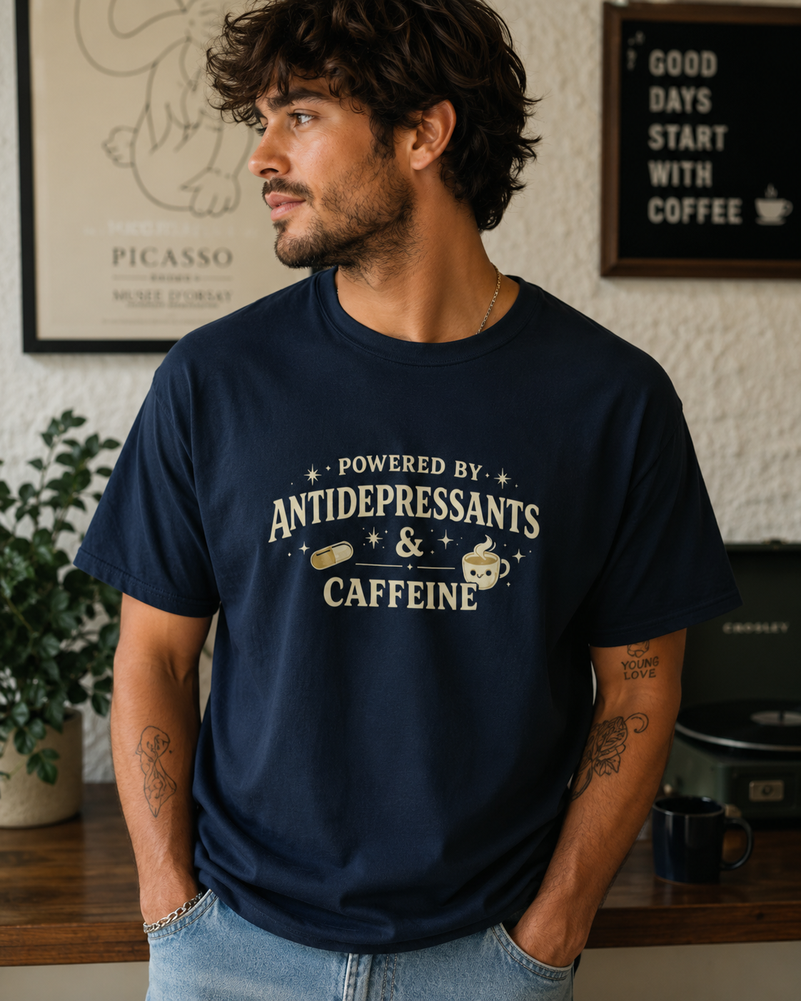 A man wearing a navy blue graphic t-shirt with a cream-colored print that reads 'POWERED BY ANTIDEPRESSANTS & CAFFEINE' with a coffee cup and pill graphic. The background shows a cozy indoor setting with framed art and a sign that says 'GOOD DAYS START WITH COFFEE'.