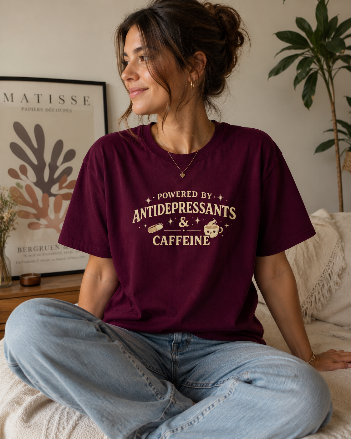 A woman sitting on a bed wearing a maroon t-shirt with a cream graphic that reads 'POWERED BY ANTIDEPRESSANTS & CAFFEINE'.