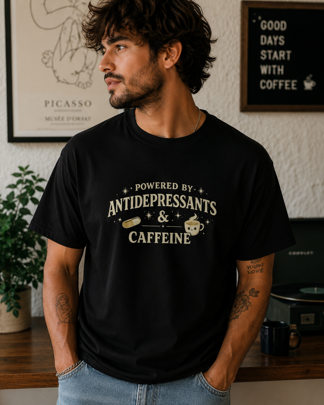 A man wearing a black graphic t-shirt with a vintage-style cream print that reads 'POWERED BY ANTIDEPRESSANTS & CAFFEINE' with coffee cup and pill illustrations. The background shows a cozy room setting with framed art and a sign.