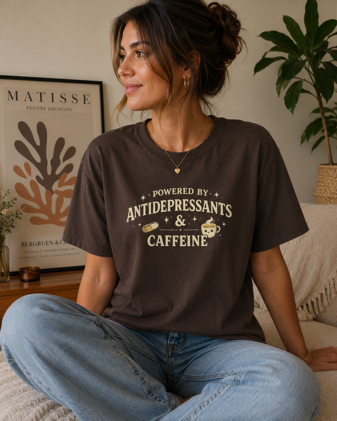 A woman sitting cross-legged on a couch wearing a dark brown graphic t-shirt that reads 'POWERED BY ANTIDEPRESSANTS & CAFFEINE' in cream lettering. The background shows a cozy living room setting with a framed Matisse print and a potted plant.