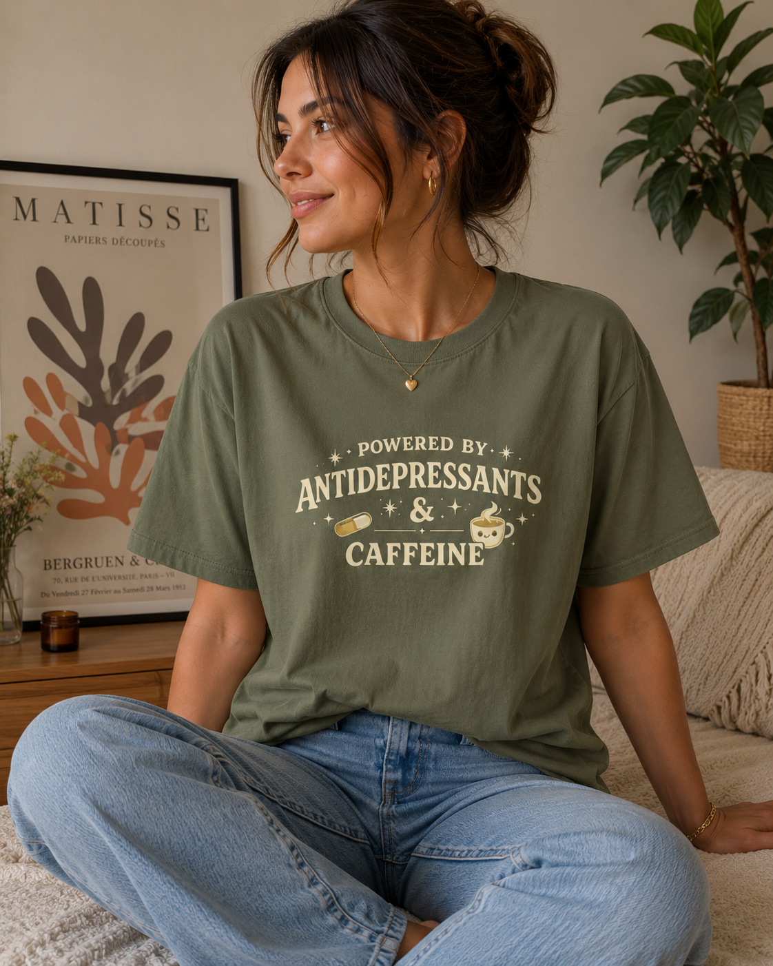 A woman sitting cross-legged on a couch wearing an olive green graphic t-shirt that reads 'POWERED BY ANTIDEPRESSANTS & CAFFEINE' with coffee cup and cookie illustrations. The background shows a cozy living room setting with a framed Matisse print and a potted plant.