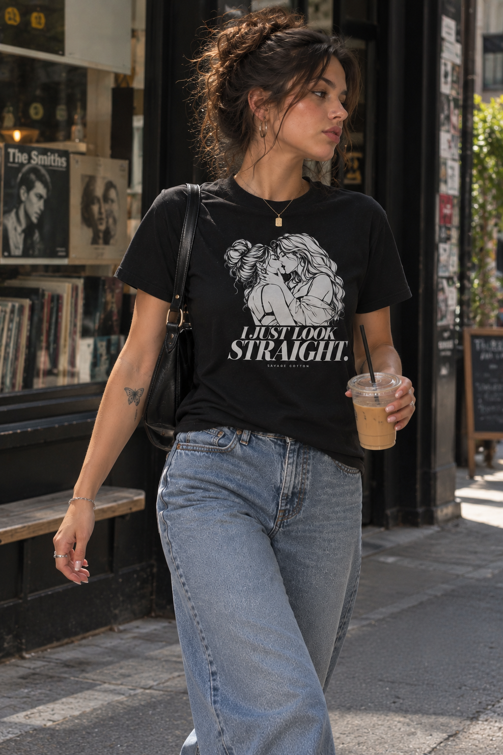 A woman walking on a city street wearing a black graphic t-shirt with a white illustration and the text 'I JUST LOOK STRAIGHT'. She is also wearing light-wash blue jeans and holding an iced coffee.