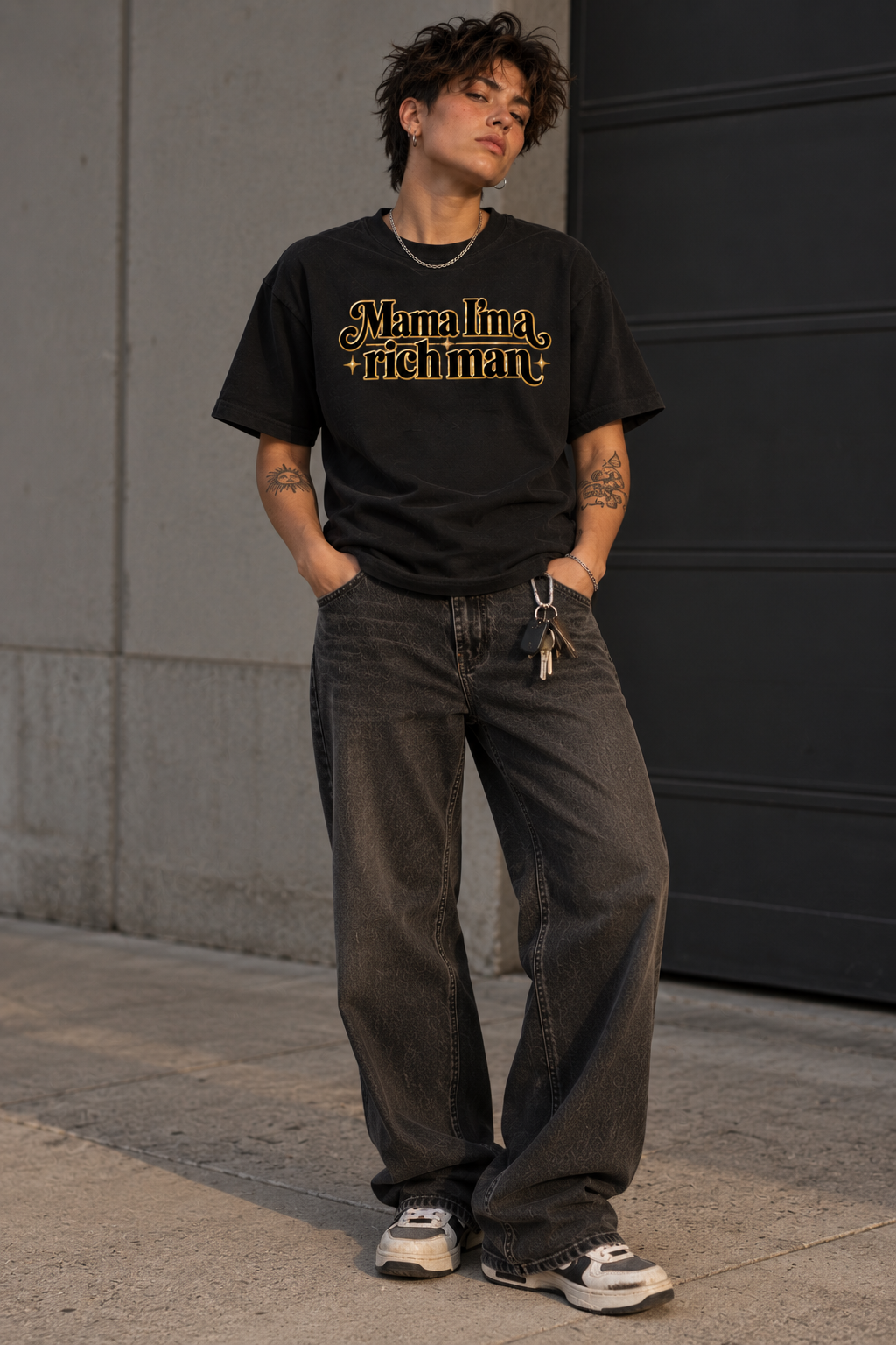 A person standing outdoors against a concrete wall and dark door, wearing a black graphic T-shirt with gold text reading 'Manalita + Richman', loose dark gray jeans, and white sneakers.