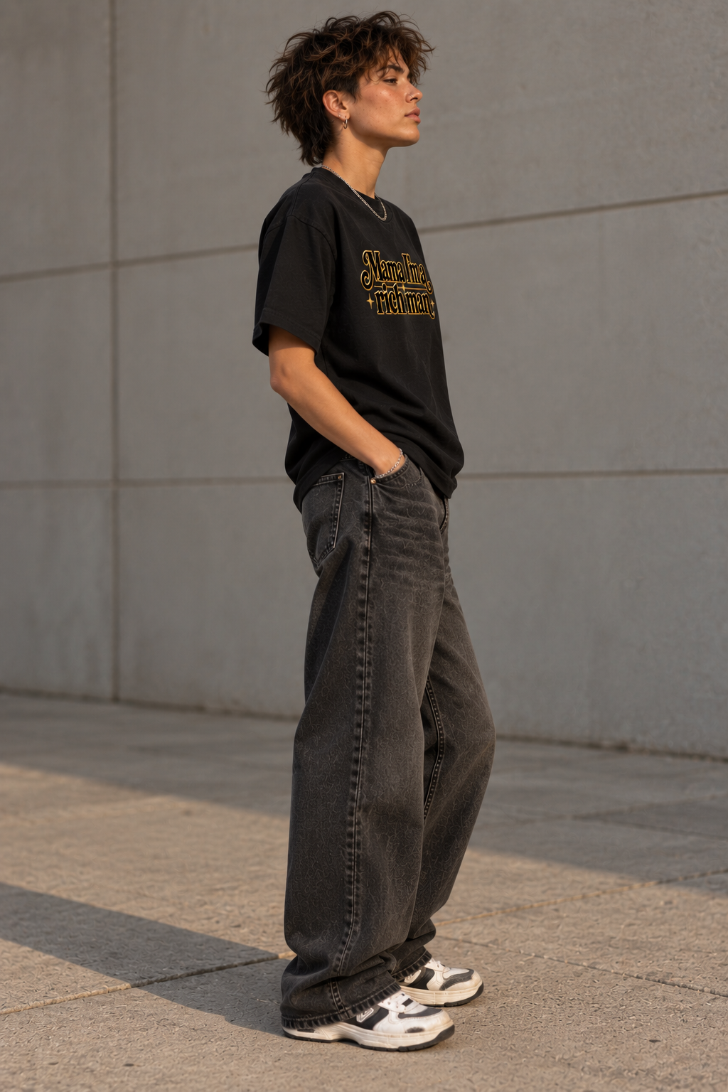 A person standing outdoors against a concrete wall, wearing a black graphic T-shirt with gold lettering, loose dark gray jeans, and white sneakers.