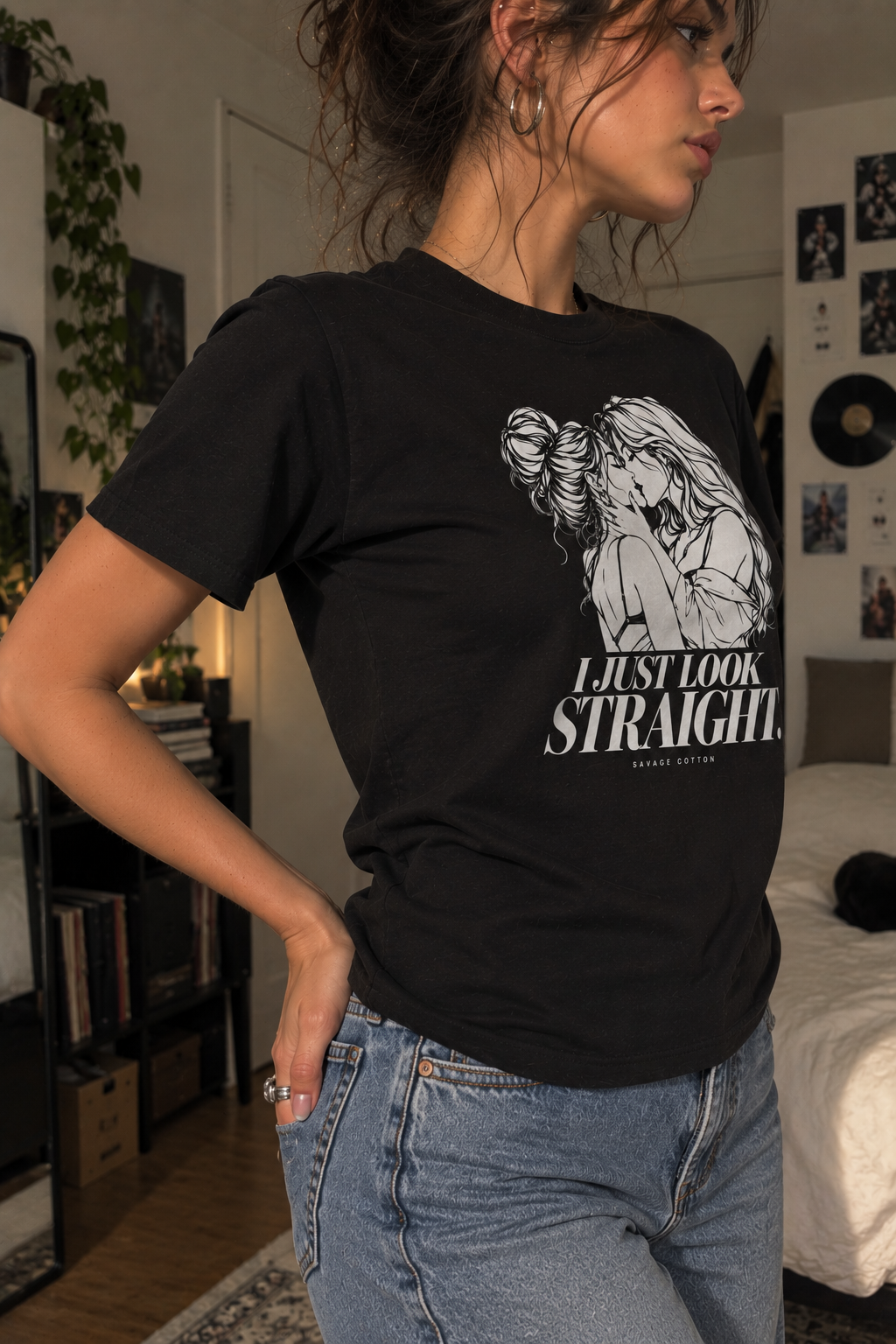 A woman wearing a black graphic t-shirt with a white illustration and the text 'I JUST LOOK STRAIGHT.' paired with light blue jeans in a bedroom setting.