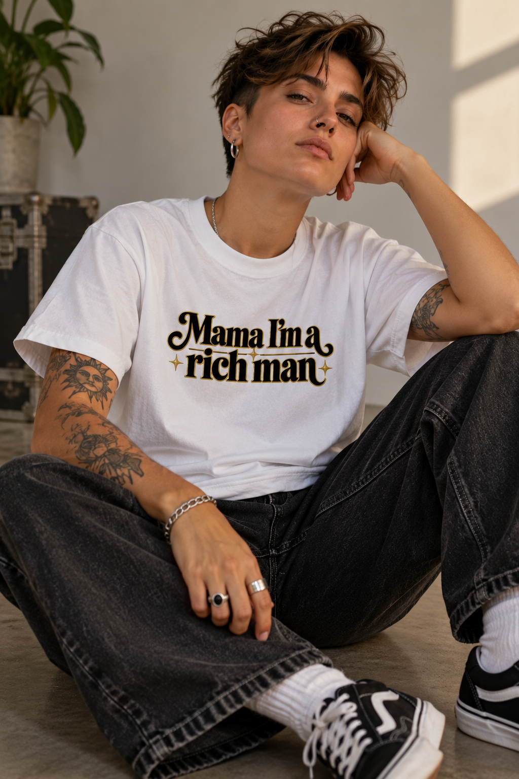 A person sitting on a floor wearing a white graphic T-shirt with the text 'Mama I'm a rich man' in black and gold, paired with dark jeans, white socks, and black sneakers.