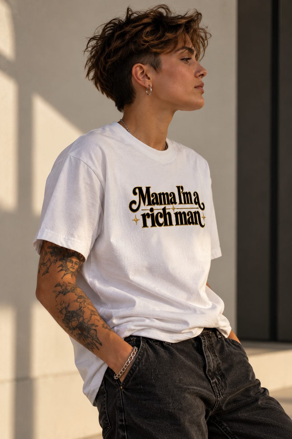 A person wearing a white graphic T-shirt with the text 'Mama I'm a, rich man' in black script, paired with dark jeans, standing against a sunlit beige wall.