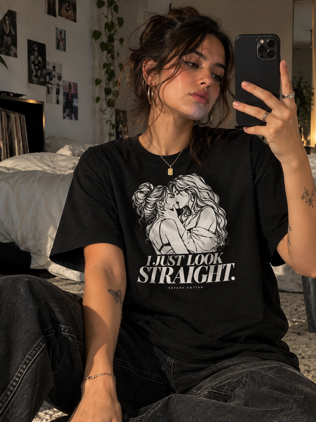 A woman takes a mirror selfie in a bedroom while wearing a black graphic t-shirt with a white illustration of a couple and the text 'I JUST LOOK STRAIGHT.' She is wearing dark gray jeans and holding a black smartphone.