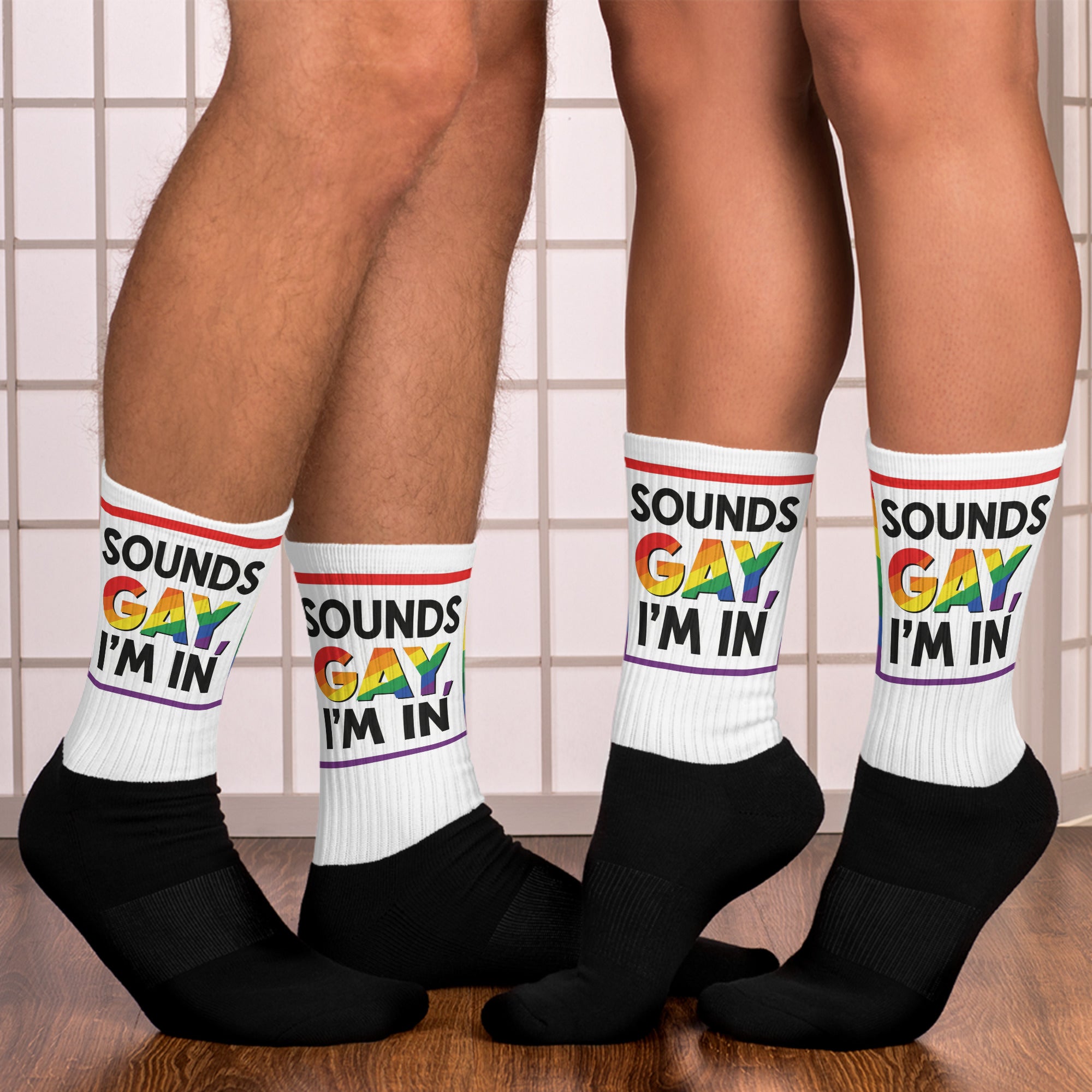 Two pairs of legs wearing socks with 'SOUNDS GAY, I'M IN' text in a rainbow and black and white color scheme.