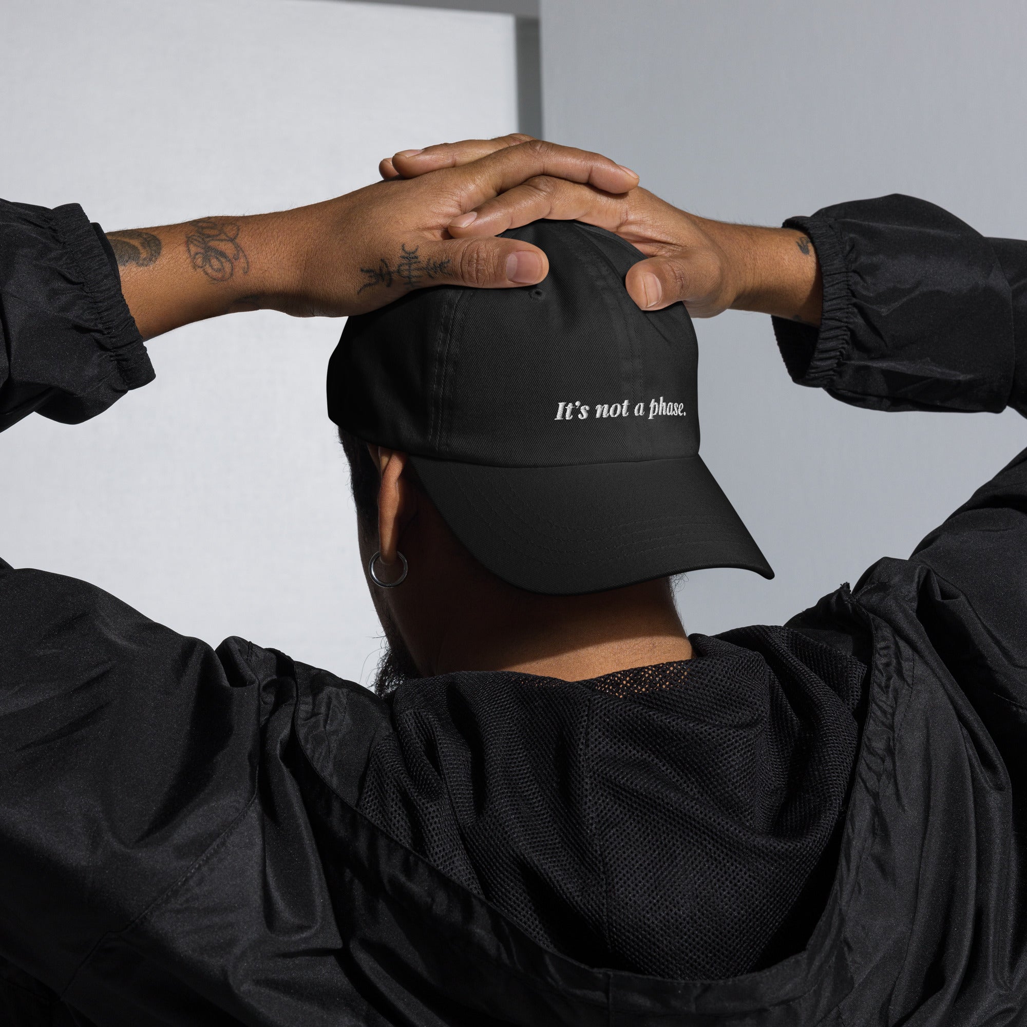 Person wearing a black cap with 'It's not a phone.' text, hands on head, plain background