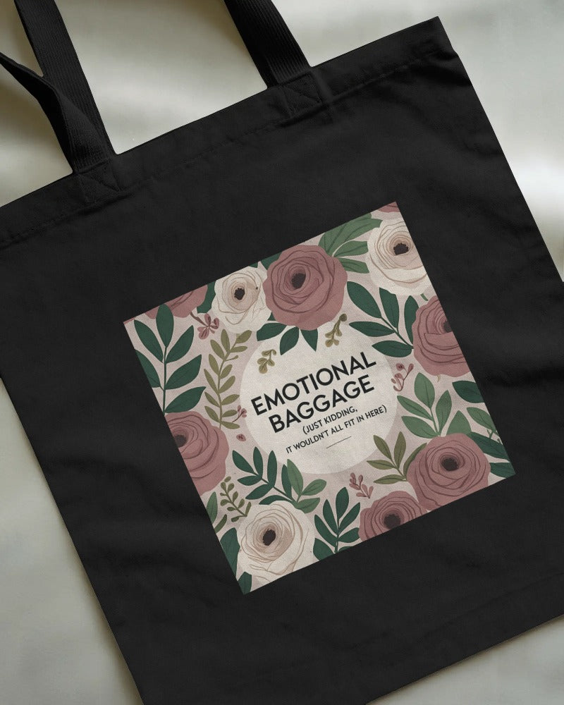 Black canvas tote bag with black text that reads “Emotional baggage… just kidding, it wouldn’t fit here” – laid flat on a wooden surface