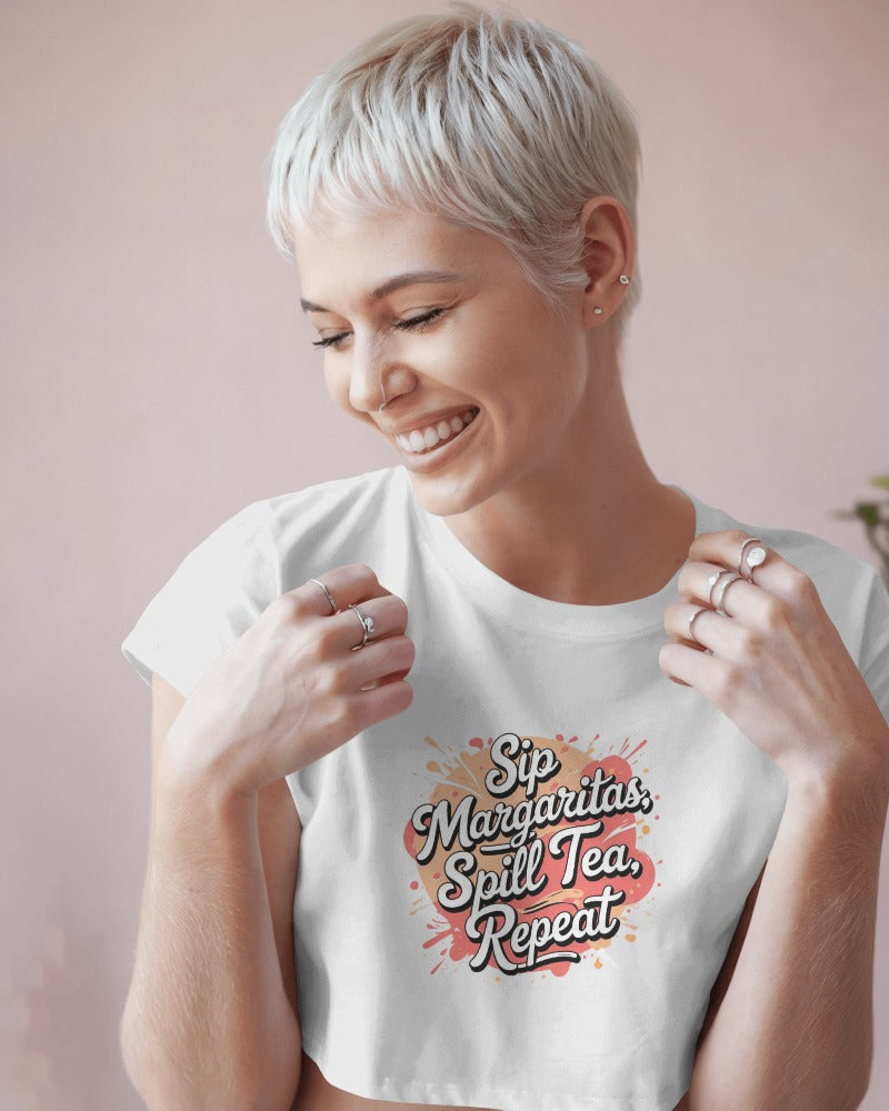 Model wearing Sip Margaritas, Spill Tea, Repeat cropped slogan T-shirt by Savage Cotton in pastel pink, styled with sunglasses and cocktail in hand