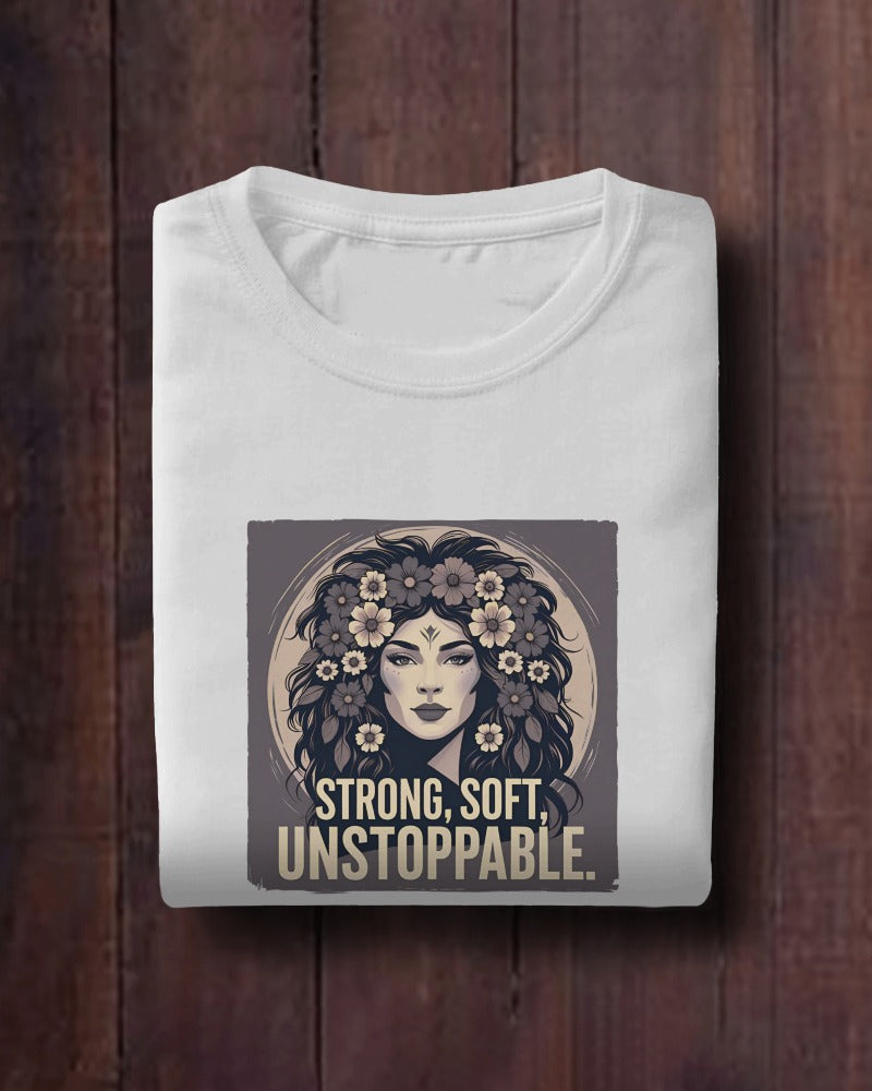 White t-shirt with a graphic design of a woman's face surrounded by flowers and text 'STRONG, SOFT, UNSTOPPABLE' on a wooden surface.