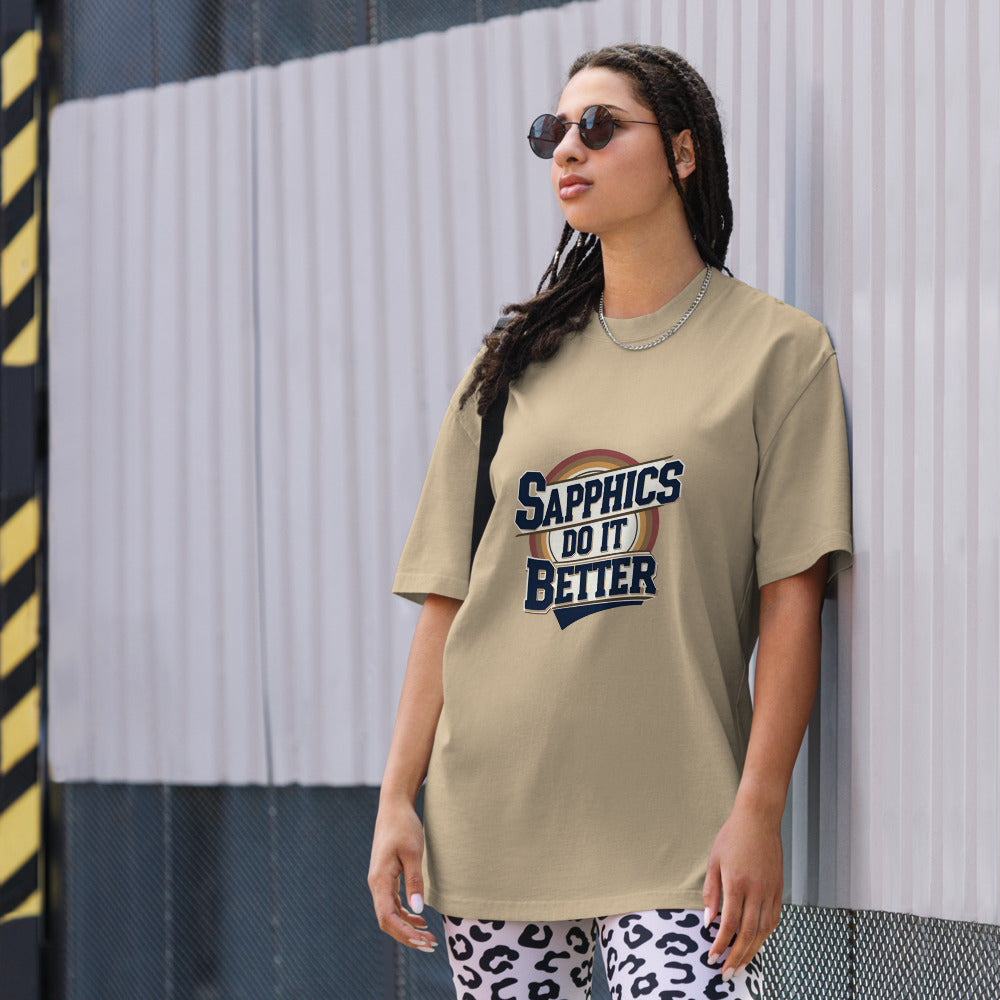 Person wearing a beige t-shirt with 'Sapphics Do It Better' text, standing against a striped wall.