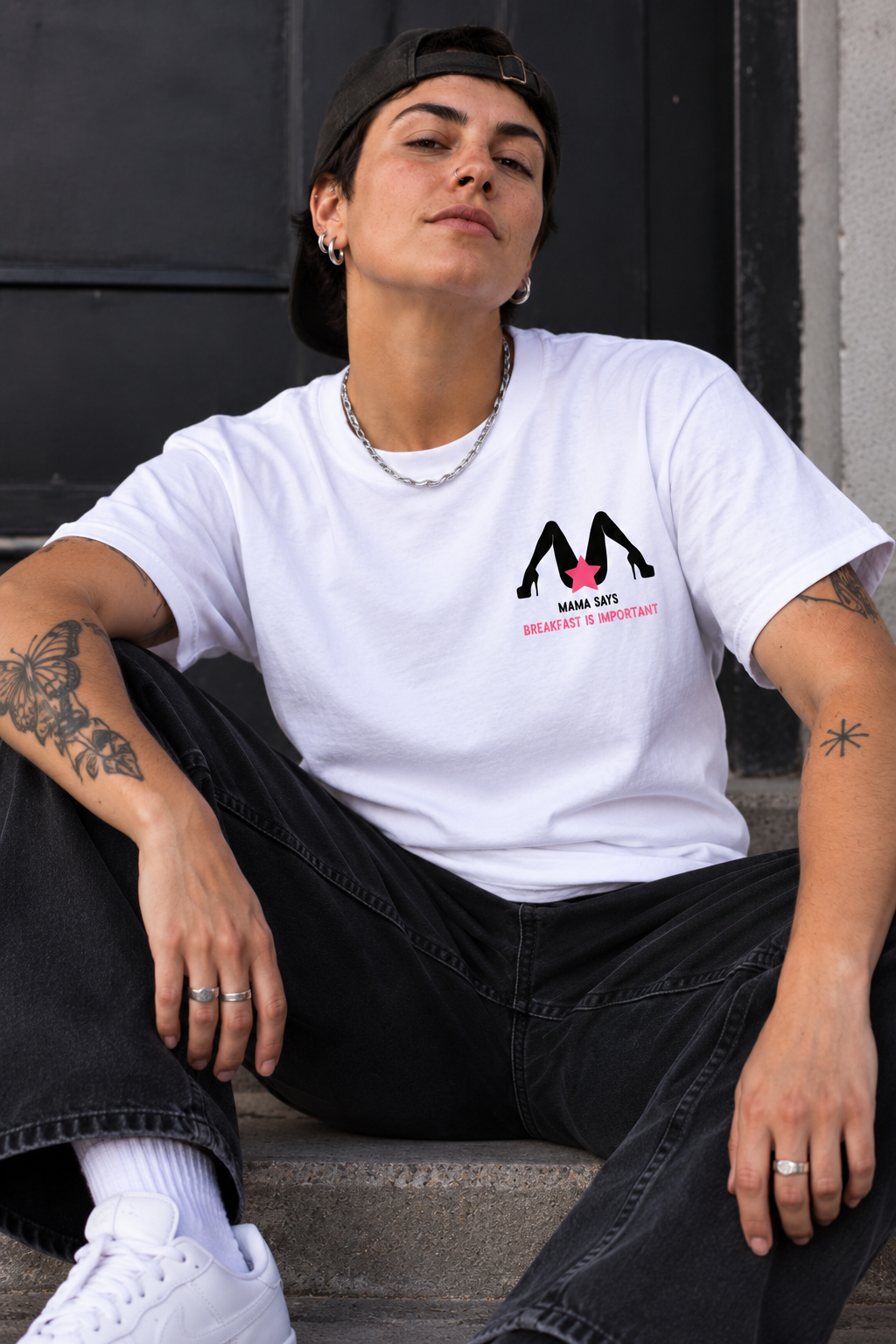 A woman wearing a white graphic t-shirt with a black and pink 'M' logo and the text 'MAMA LIPS' and 'BREATHE & SMILE' sitting on concrete steps.