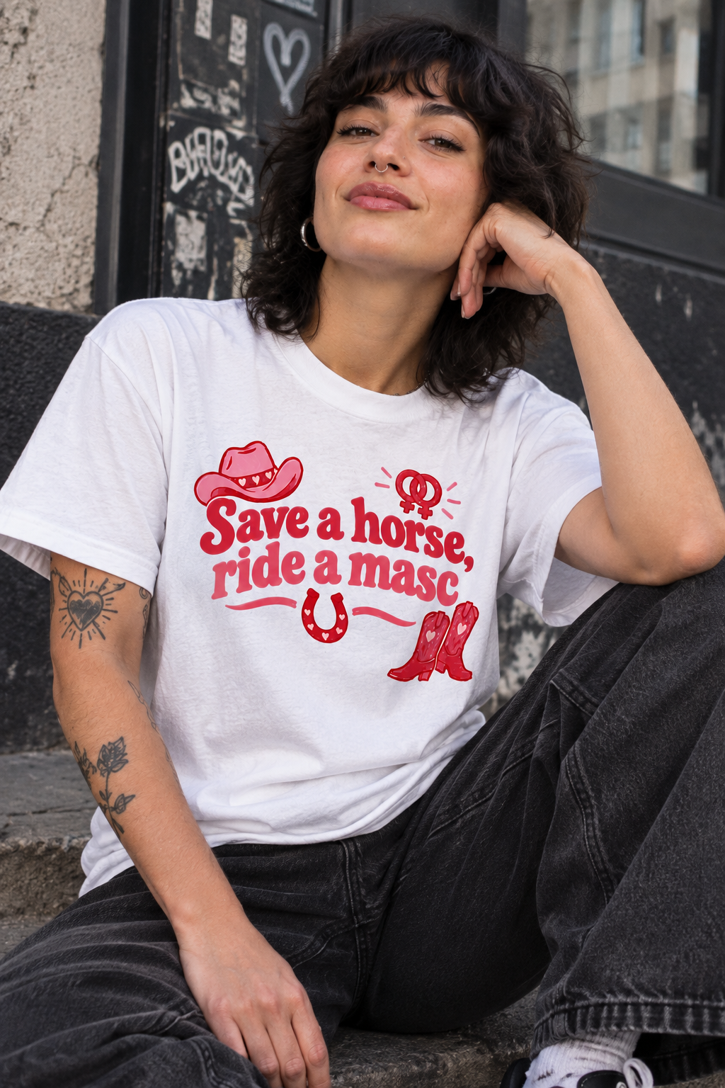 A woman wearing a white graphic t-shirt with a red western-themed print that reads 'Save a horse, ride a masc'. She is sitting outdoors on a stone ledge next to a graffiti-covered wall.