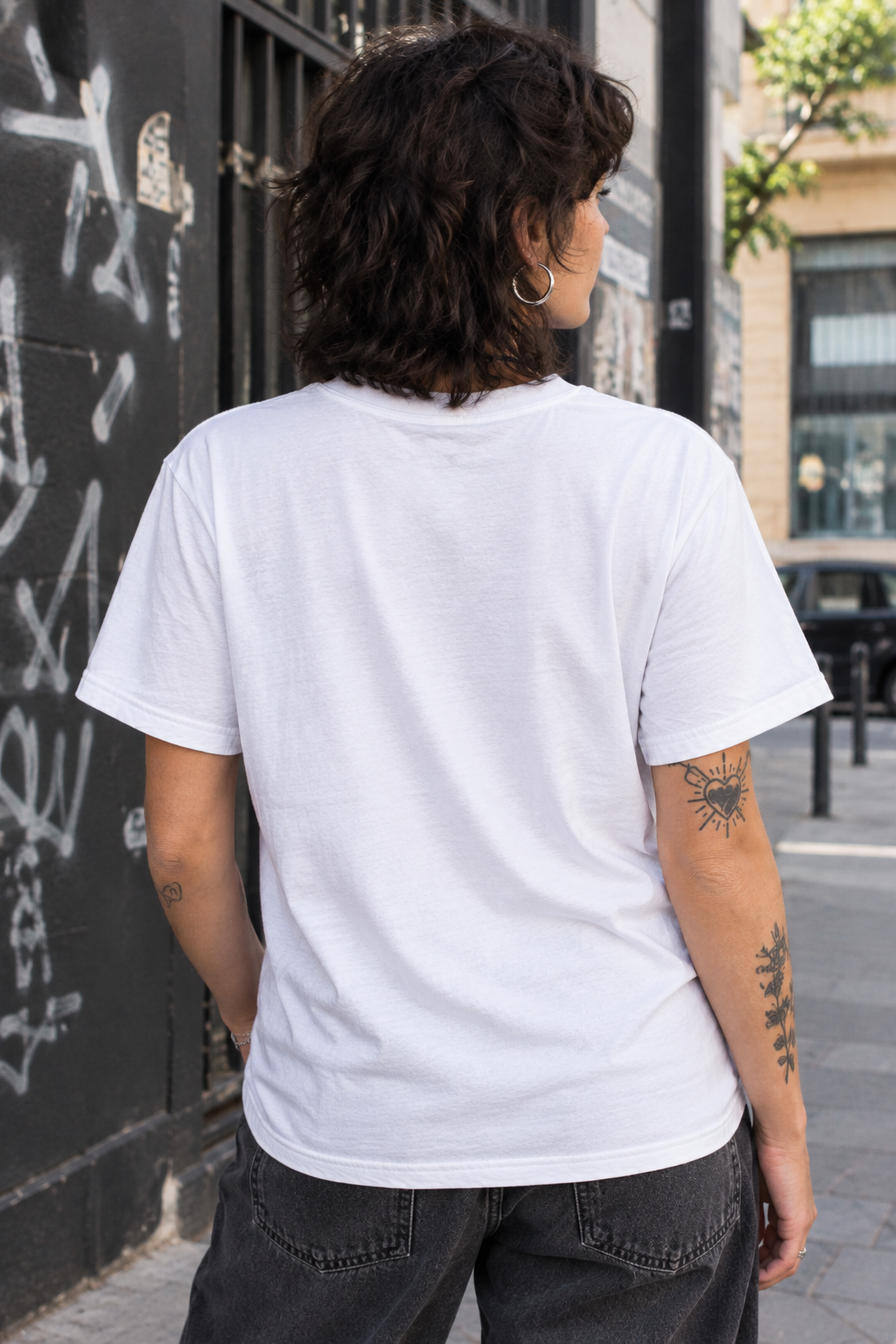 A person viewed from behind wearing a plain white short-sleeve crewneck t-shirt and dark gray jeans, standing outdoors on a city street next to a graffiti-covered wall.