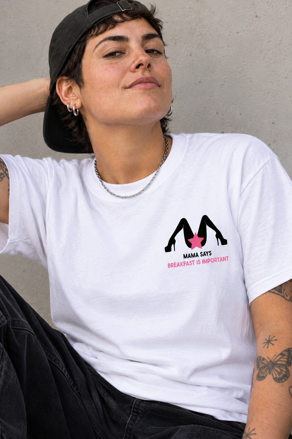 A person wearing a white graphic T-shirt with a black high-heel logo and pink text reading 'MAMA SAYS BREAKFAST IS IMPORTANT', paired with a black cap and dark pants, posed against a concrete wall.