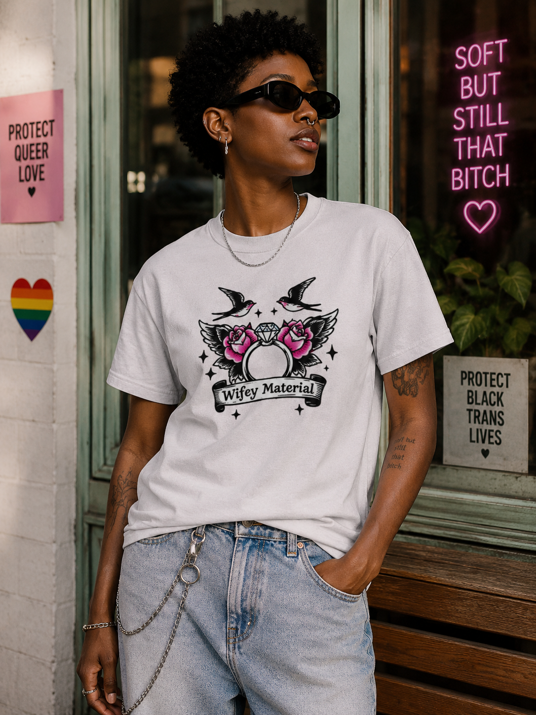 A person wearing a white graphic T-shirt with a black and pink winged egg design and the text 'Wifey Material,' paired with light blue jeans, standing outdoors in front of a storefront with pride-themed signs.