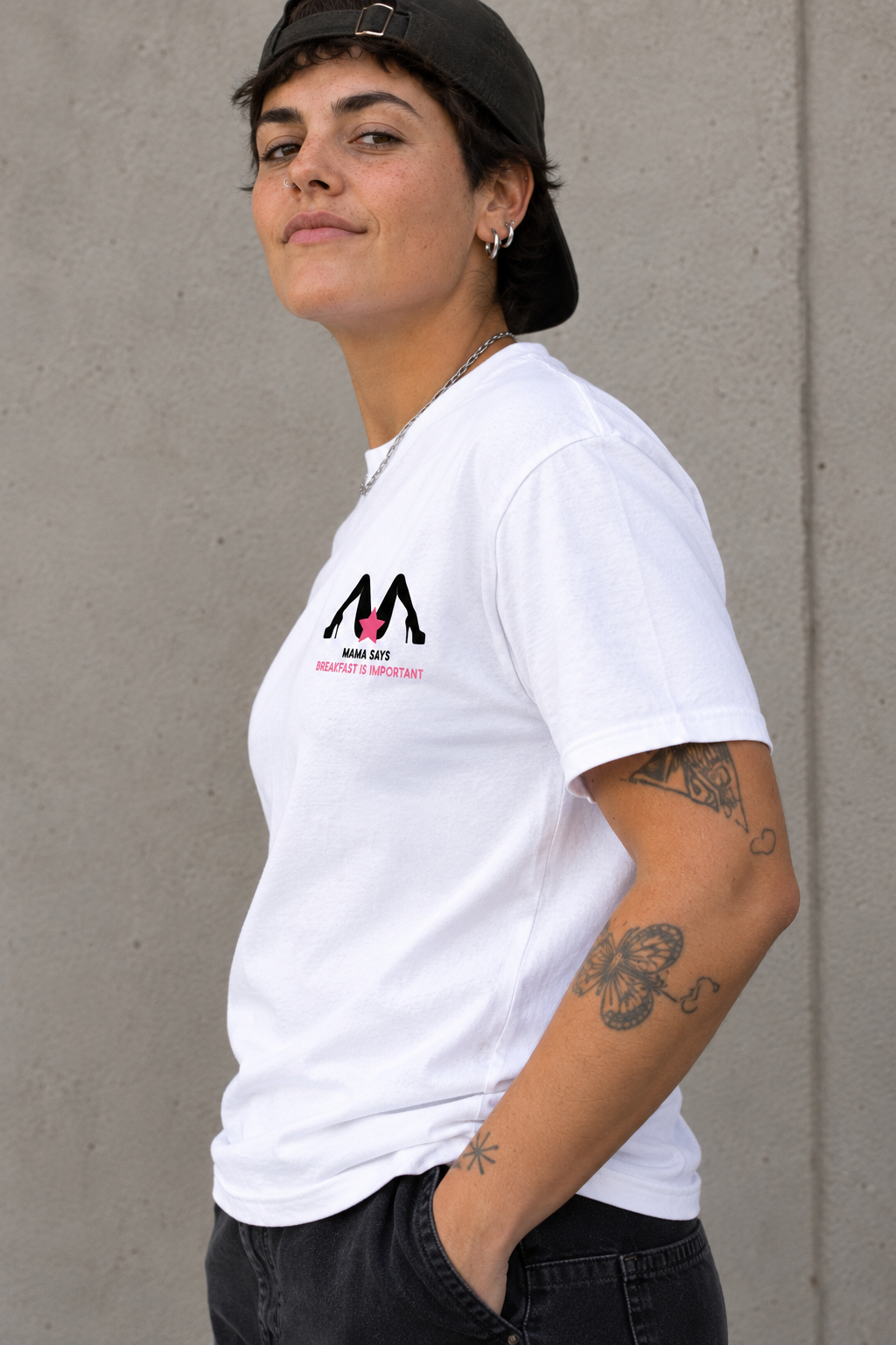 A woman wearing a white short-sleeve t-shirt with a black and pink 'M' logo on the left chest, paired with a black baseball cap and dark jeans, standing against a concrete wall.