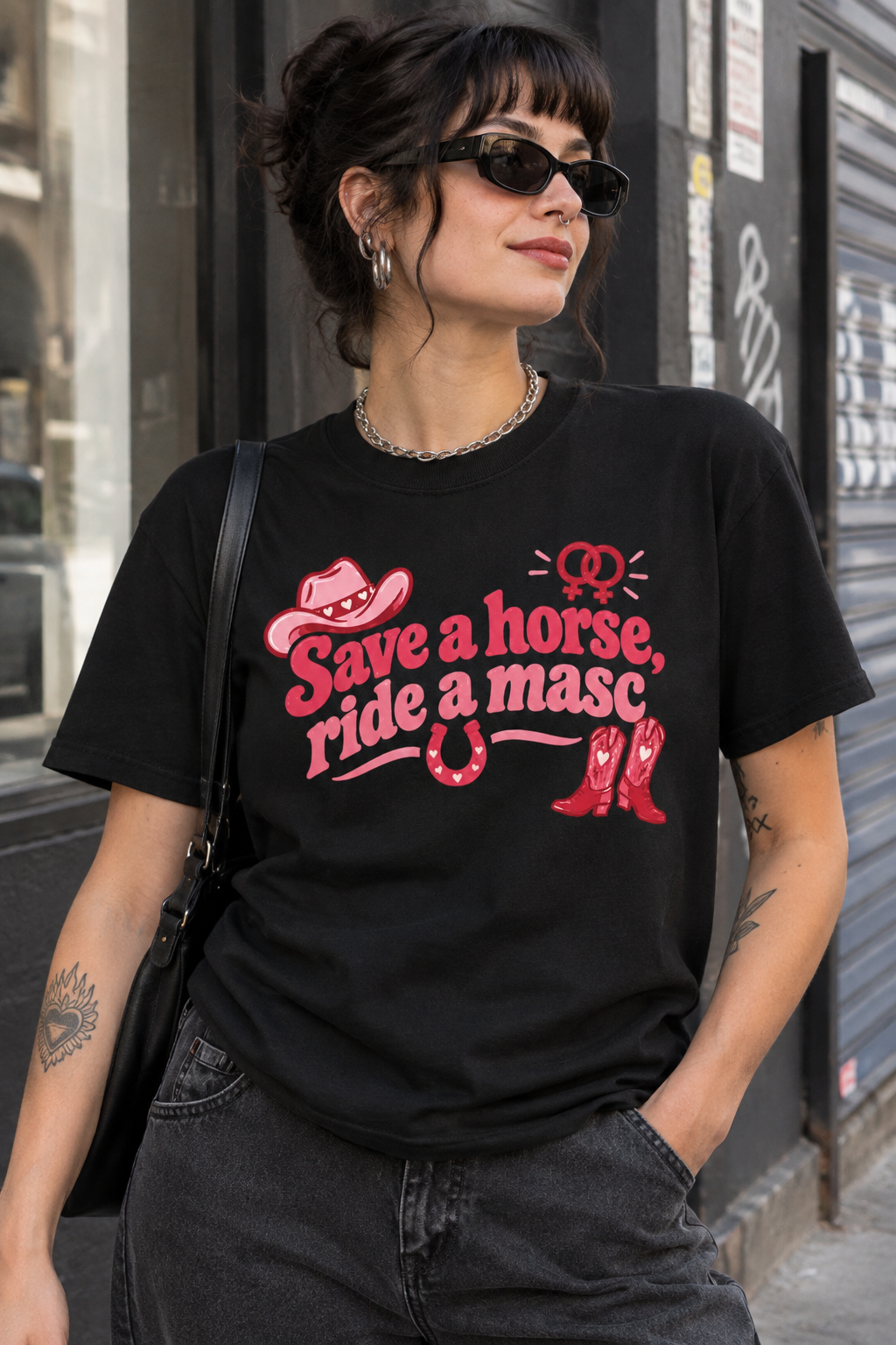 A woman wearing a black graphic t-shirt with a red western-themed print that reads 'Save a horse, ride a masc'. She is wearing dark sunglasses, a silver chain necklace, and dark gray jeans, carrying a black shoulder bag in an outdoor urban setting.