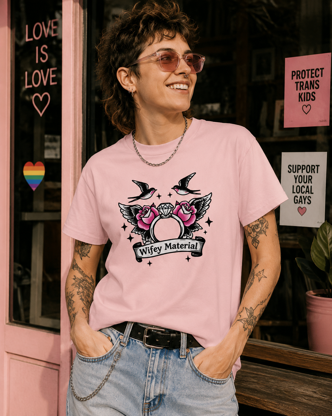 A person wearing a light pink graphic T-shirt with a black, white, and pink 'Wifey Material' design, paired with light blue jeans, standing in front of a pink storefront with supportive signs.