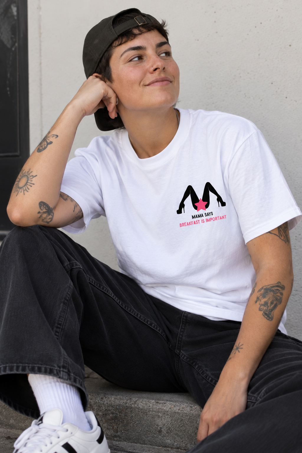 A person sitting outdoors wearing a white graphic T-shirt with a black and pink 'M' logo and the text 'MUMBA SHIT IS IMPORTANT', paired with black jeans, white socks, and white sneakers.