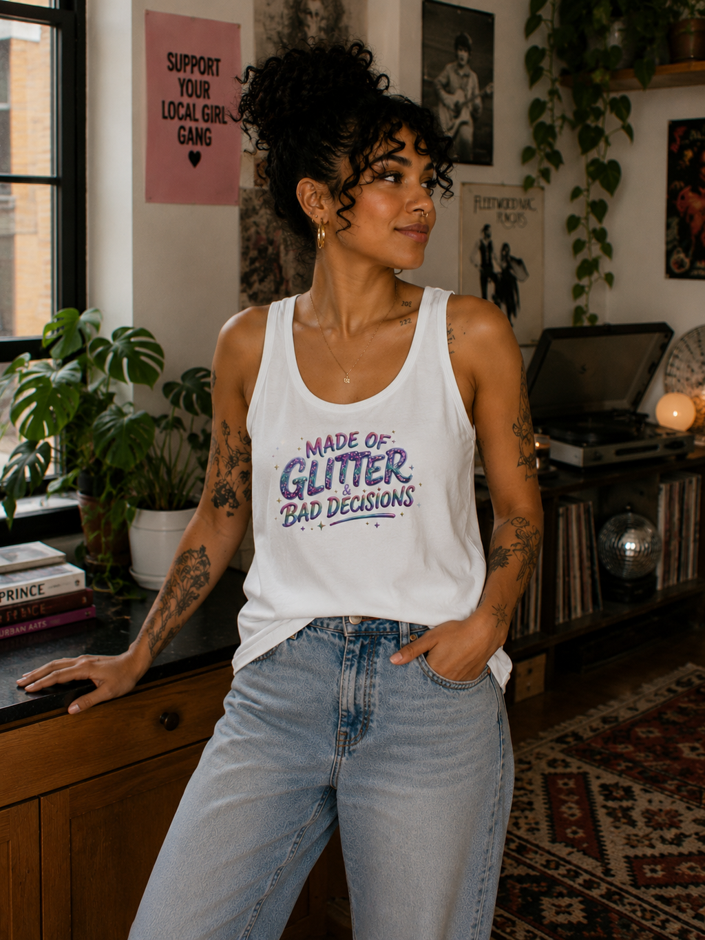 A woman with curly hair stands in a cozy, decorated room wearing a white tank top with the text 'MADE OF GLITTER BAD DECISIONS' in purple and blue. She is also wearing light-wash blue jeans.