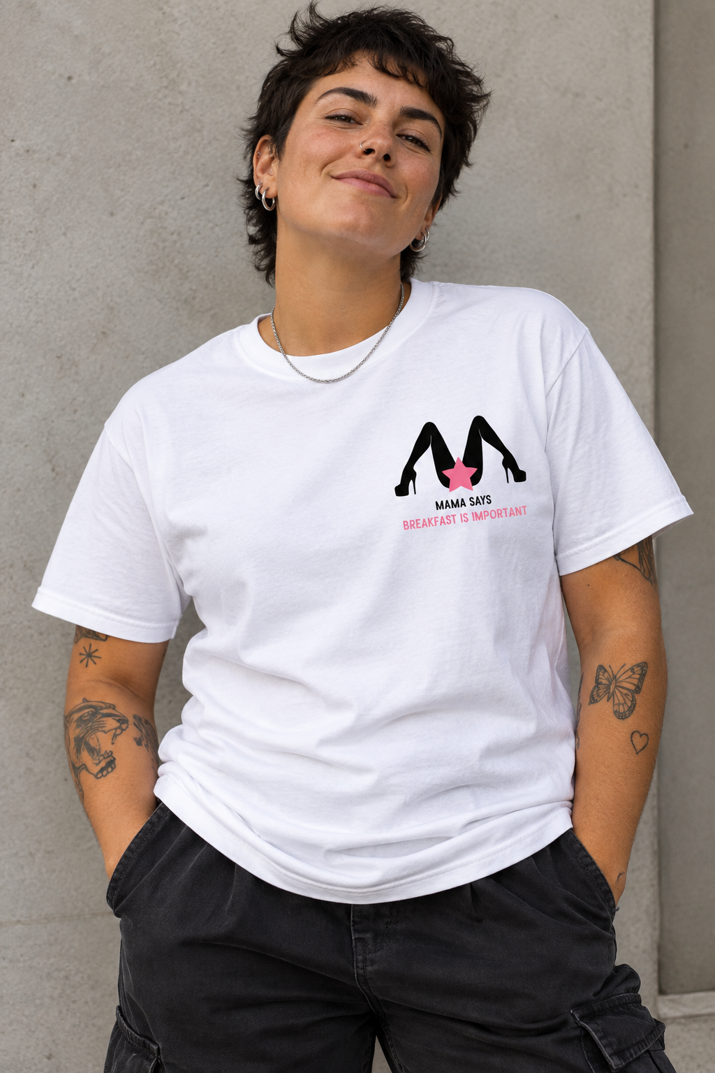 A woman wearing a white graphic t-shirt with a black and pink high-heel logo and the text 'MAKE LOVE GREAT AGAIN' on the chest, paired with black cargo pants, standing against a textured gray concrete wall.
