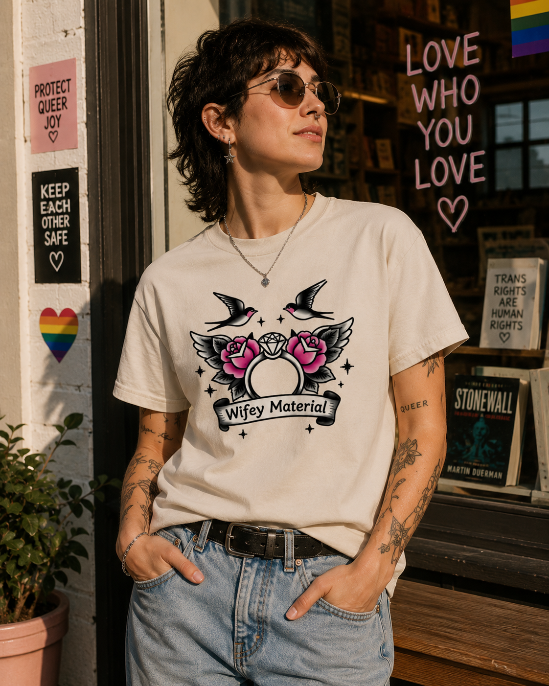 A woman wearing a cream-colored graphic t-shirt with a 'Wifey Material' design featuring two birds and pink roses, paired with light-wash blue jeans, standing in front of a storefront window.