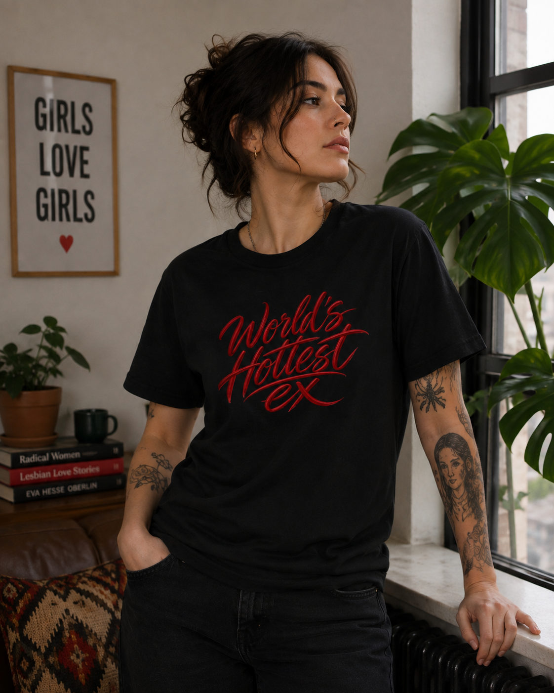 A woman wearing a black graphic t-shirt with red text that reads 'World's Hottest EX', standing in a room with a window, plants, and a framed sign on the wall.