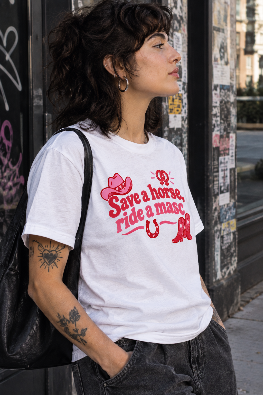 A woman wearing a white graphic t-shirt with a red western-themed print that reads 'Save a horse, ride a masc' alongside cowboy hat, boots, and horseshoe illustrations, photographed in an outdoor urban setting.
