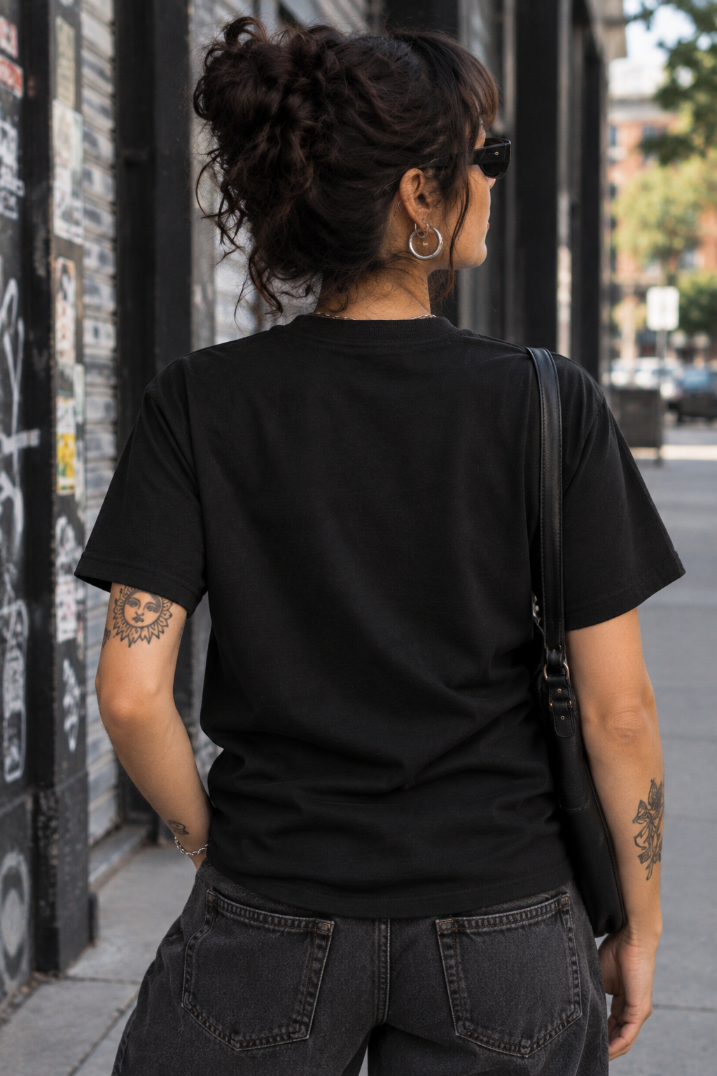 A woman viewed from behind wearing a plain black short-sleeve t-shirt and dark gray jeans, standing on a city sidewalk next to a graffiti-covered wall.