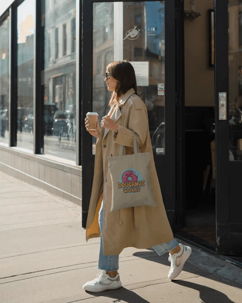 Woman in a trench coat holding a coffee cup and a tote bag with a logo, standing on a sidewalk.