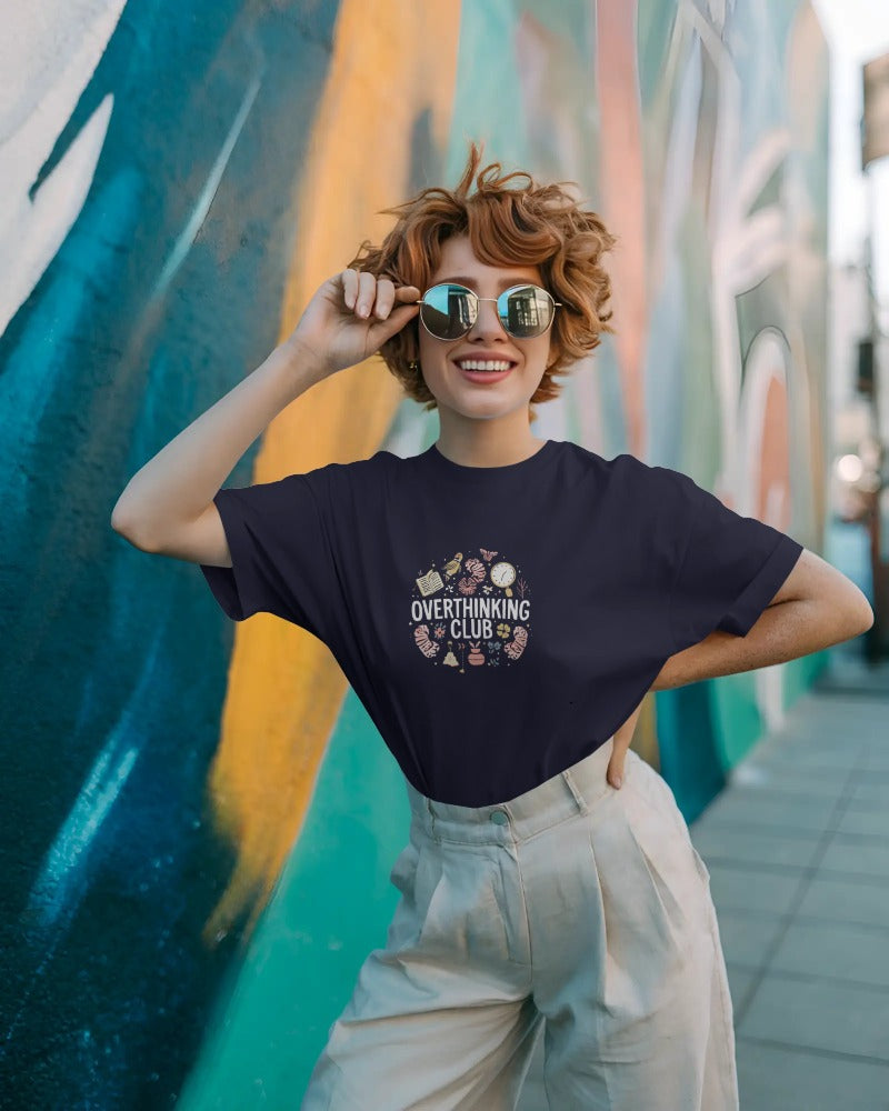 Woman wearing a navy blue t-shirt with 'Overthinking Club' design in front of a colorful mural.