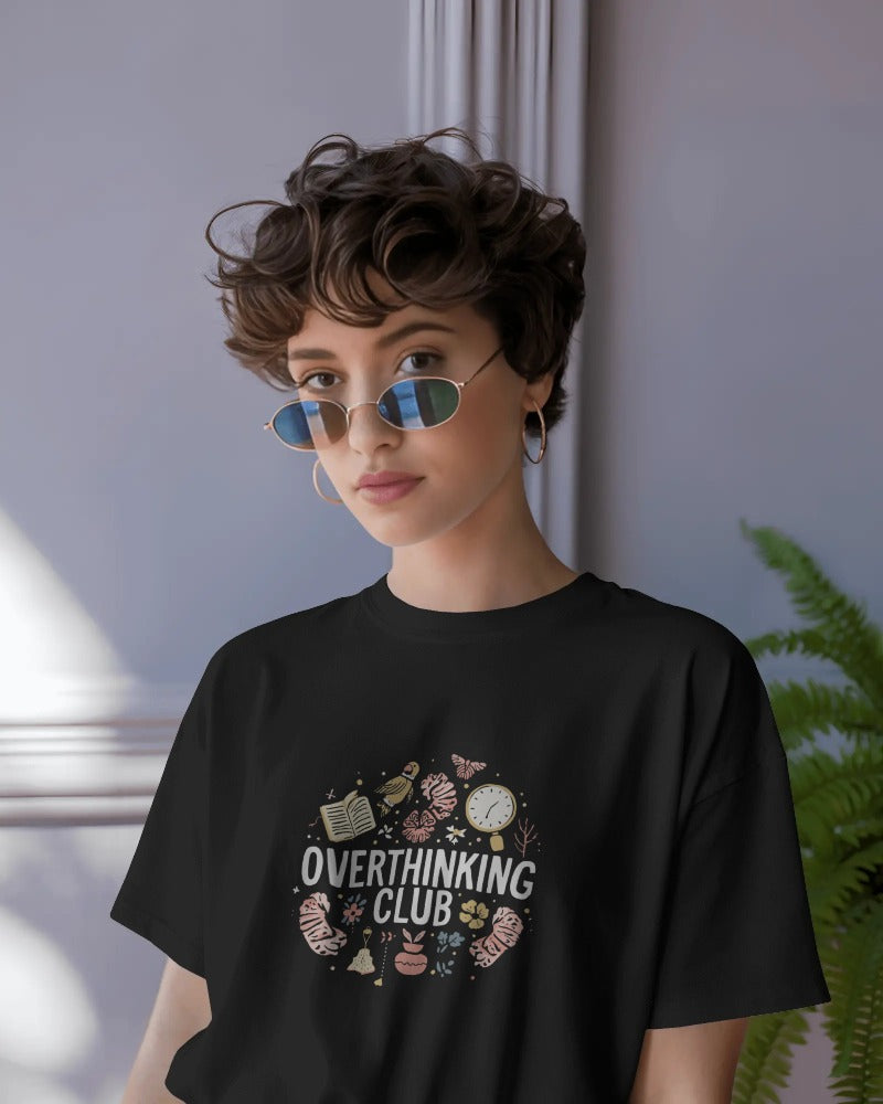 Person wearing a black t-shirt with 'Overthinking Club' design, standing indoors.