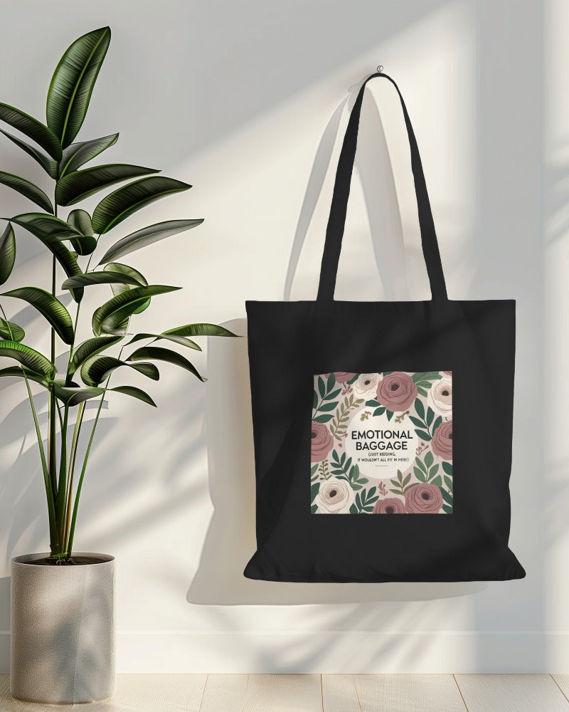 Black canvas tote bag with black text that reads “Emotional baggage… just kidding, it wouldn’t fit here” – laid flat on a wooden surface