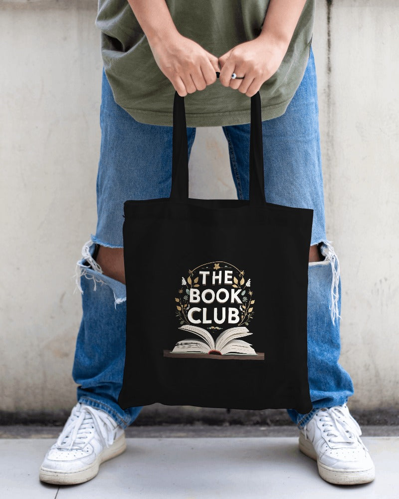 Person holding a black tote bag with 'The Book Club' design against a neutral background