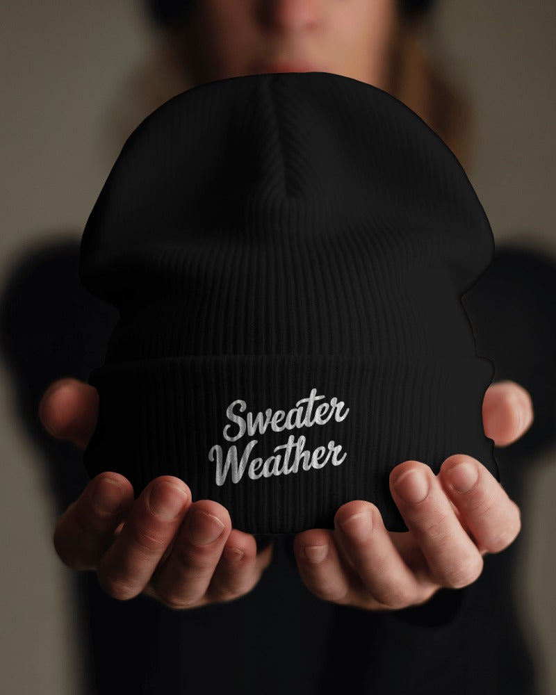 Black beanie with 'Sweater Weather' text held by hands against a blurred background