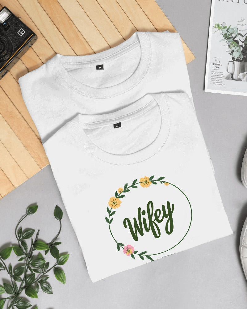 White t-shirt with 'Wifey' design on a gray surface with a plant and camera in the background