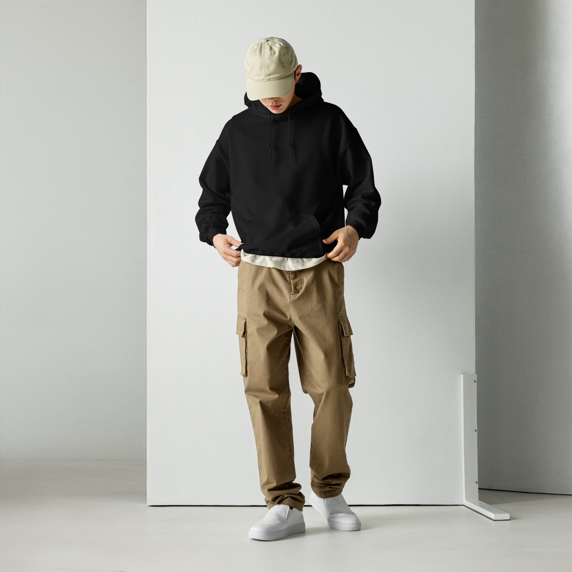 Person wearing a black hoodie, beige cap, brown cargo pants, and white shoes against a plain background