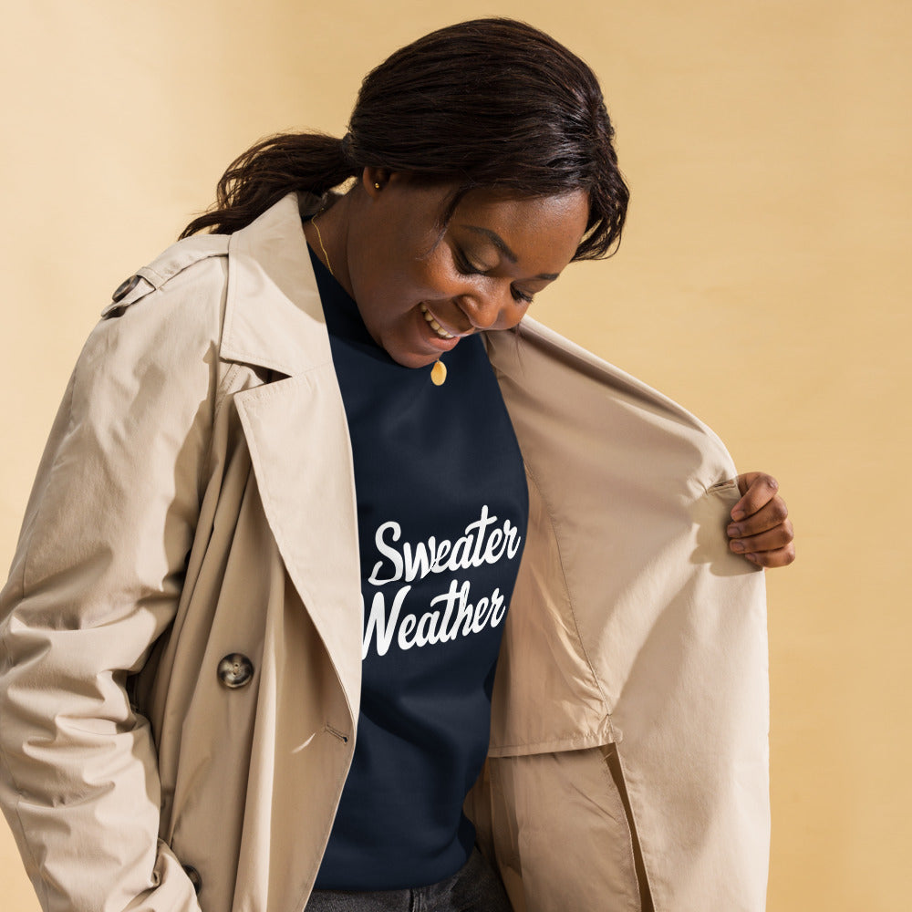 Woman wearing a navy sweatshirt with 'Sweater Weather' text, holding a beige trench coat against a beige background.